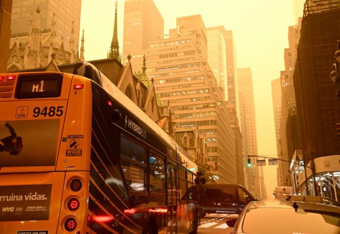 A line of traffic stretches down a Manhattan street, tower blocks rising on either side. The visibility is poor as smoke pollutes the air, turned orange by the sun.