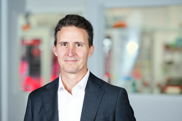 Portrait of HW Vassen in a suit and shirt looking directly at the camera, with a blurred office background