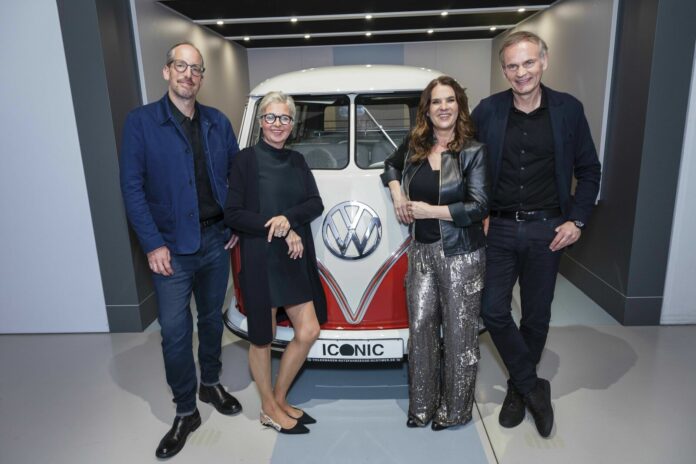 Four people stand smiling next to a classic Volkswagen bus in a showroom under the "ICONIC" theme.