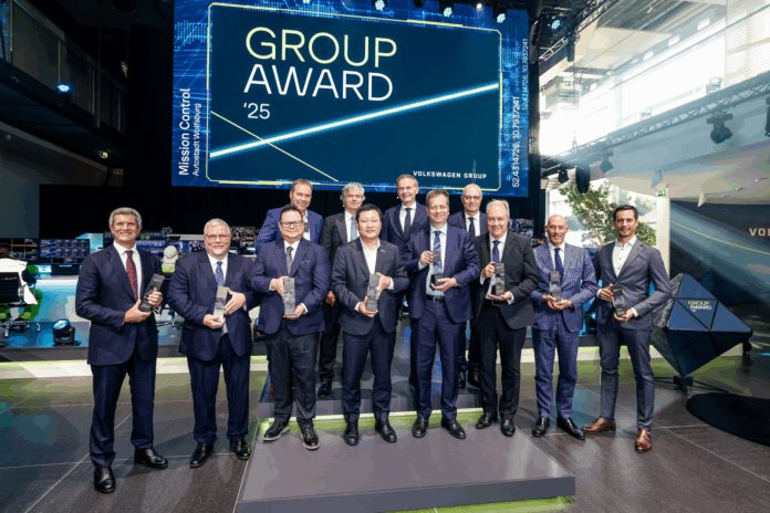Group of eleven men in suits holding trophies at the Volkswagen Group Award ceremony 2025. In the background, a large screen displays “GROUP AWARD '25” and “Volkswagen Group”.