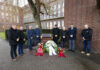 Internationaler Holocaust-Gedenktag: Volkswagen Group erinnert an die Befreiung des Konzentrationslagers Auschwitz vor 80 Jahren und gedenkt der Opfer des Holocaust An outdoor memorial event where several individuals stand in a row in front of a monument, surrounded by red and white floral wreaths. The participants bow their heads in a moment of silence. Brick buildings and a tree-lined area are visible in the background.