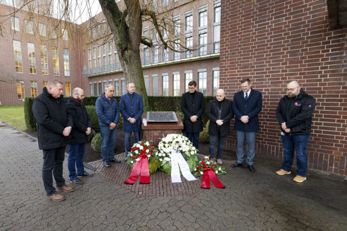 An outdoor memorial event where several individuals stand in a row in front of a monument, surrounded by red and white floral wreaths. The participants bow their heads in a moment of silence. Brick buildings and a tree-lined area are visible in the background.